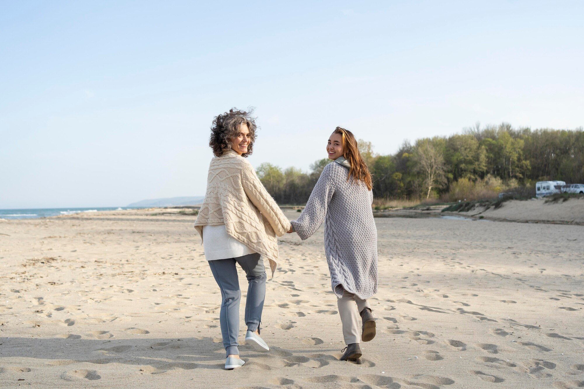 Mãe e filha de mãos dadas caminhando na praia e sorrindo para a câmera