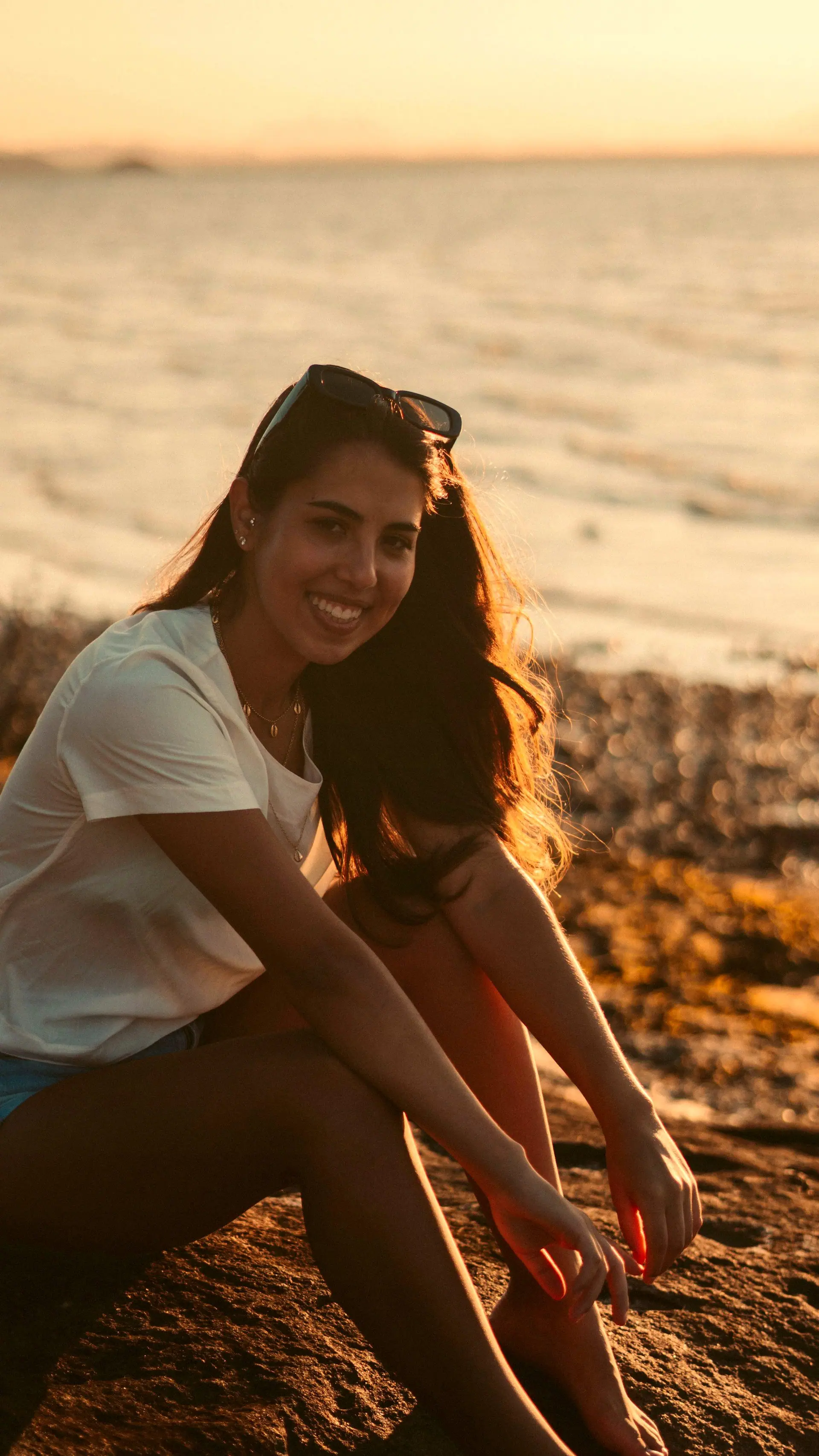 Mulher sorrindo sentada na areia, iluminada pelo sol do fim de tarde
