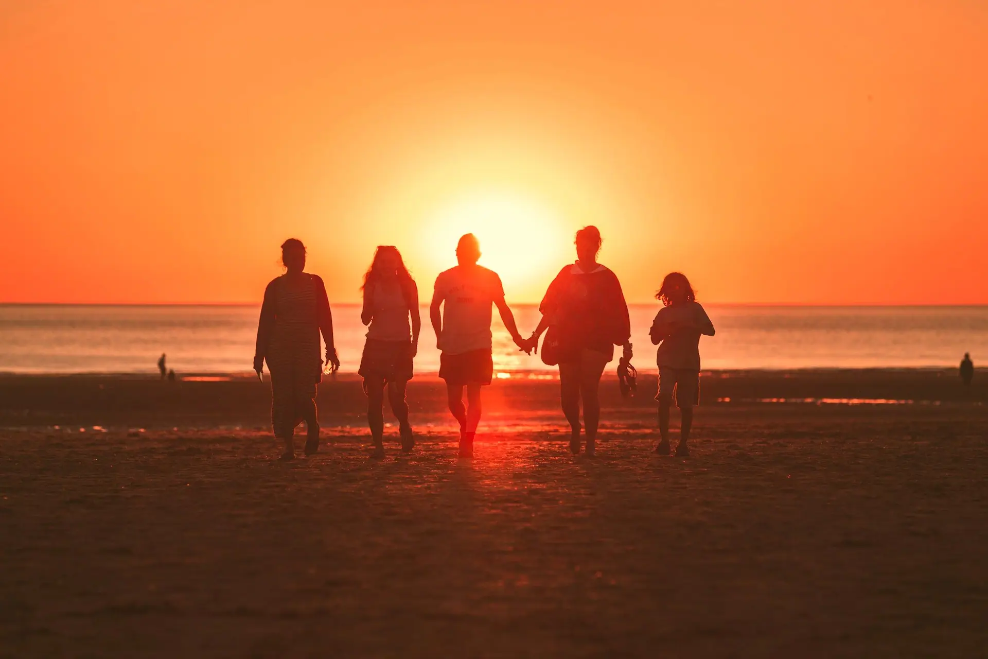 Família de cinco pessoas caminhando de mãos dadas na praia em direção ao pôr do sol