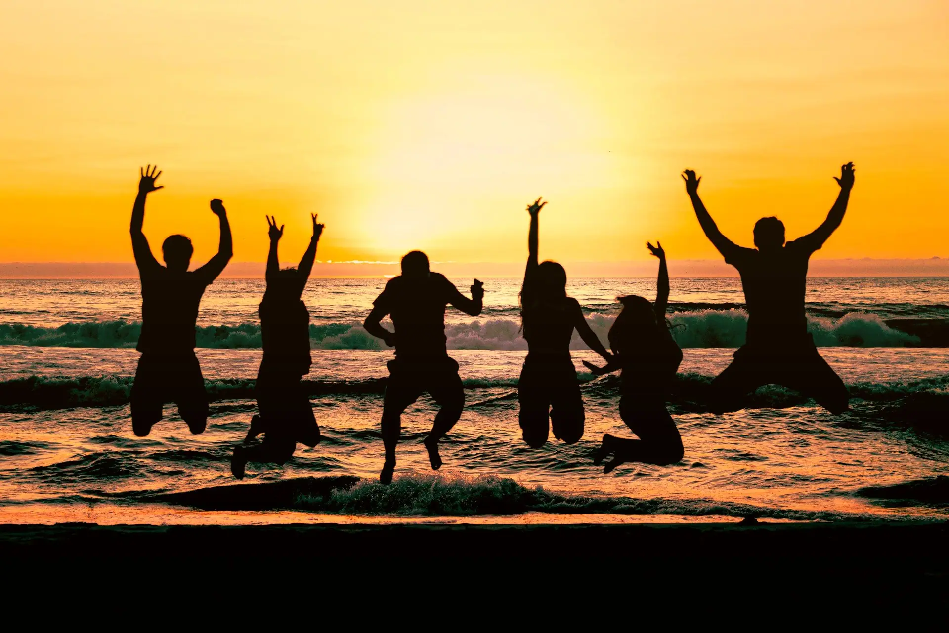 Grupo de amigos pulando juntos na beira do mar contra o sol do fim de tarde