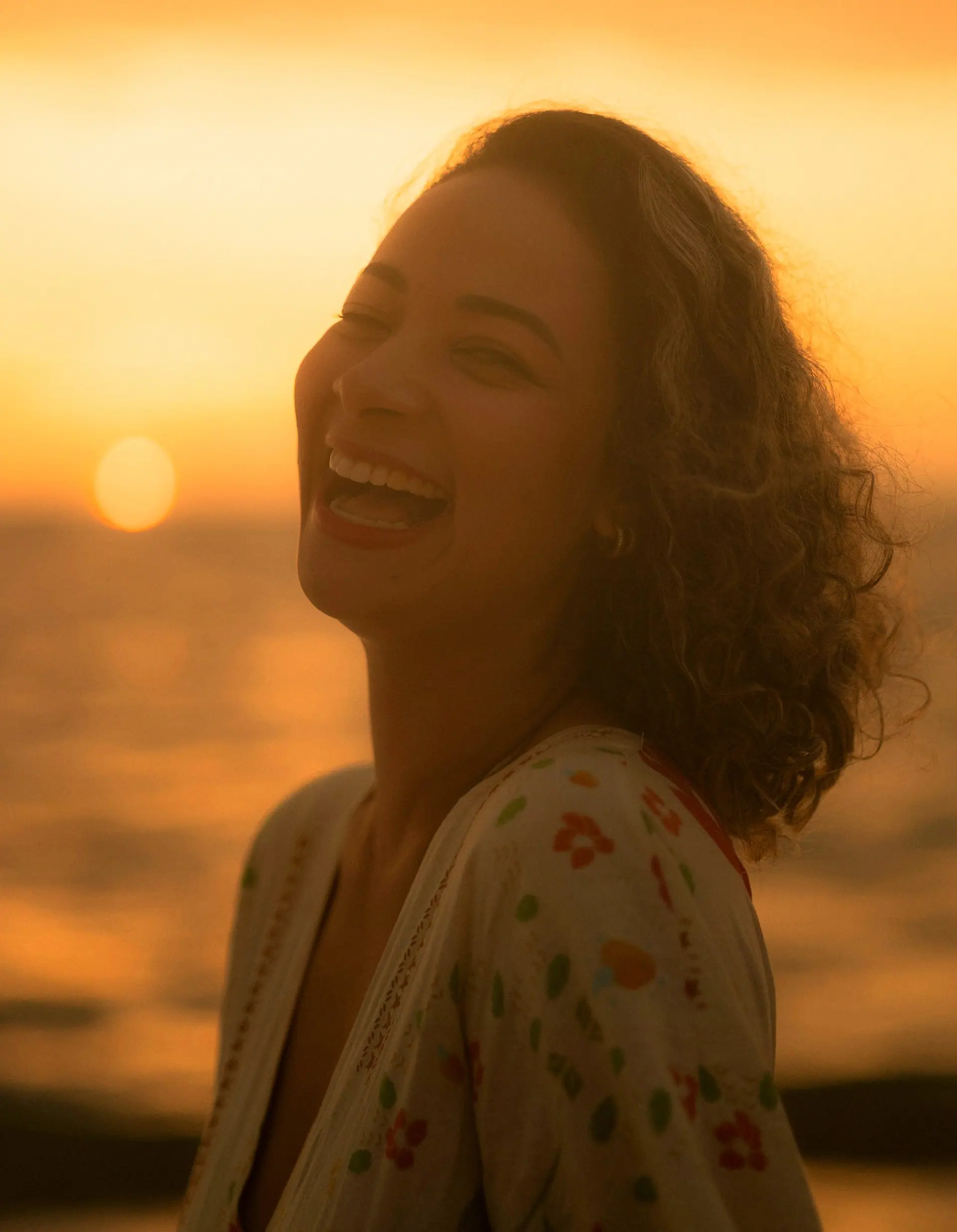 Mulher de cabelo cacheado gargalhando na praia ao pôr do sol