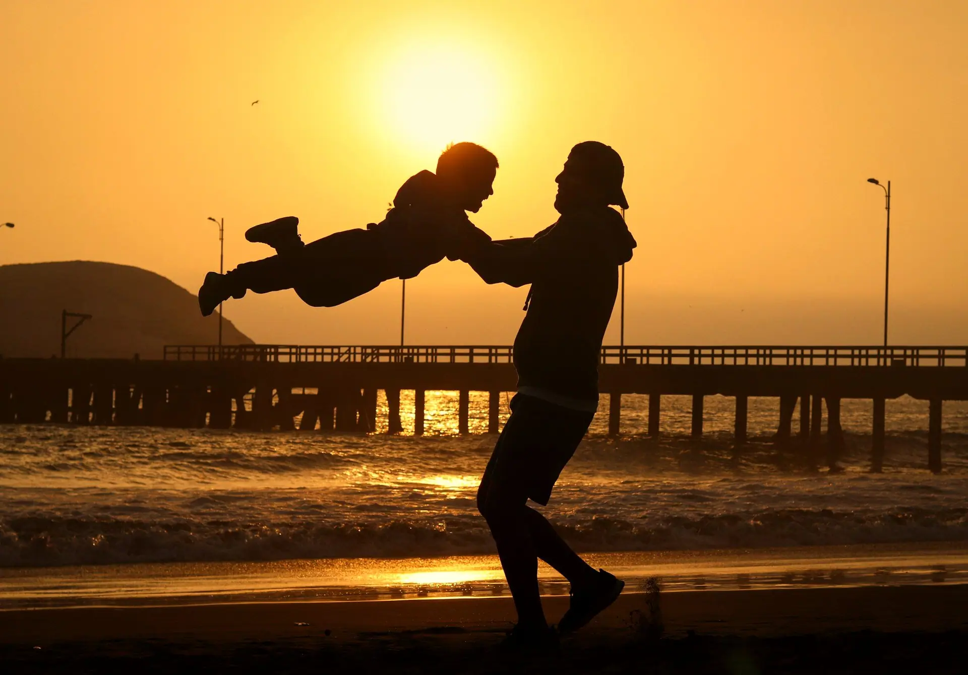 Pai brincando de aviãozinho com o filho na praia contra o sol do entardecer