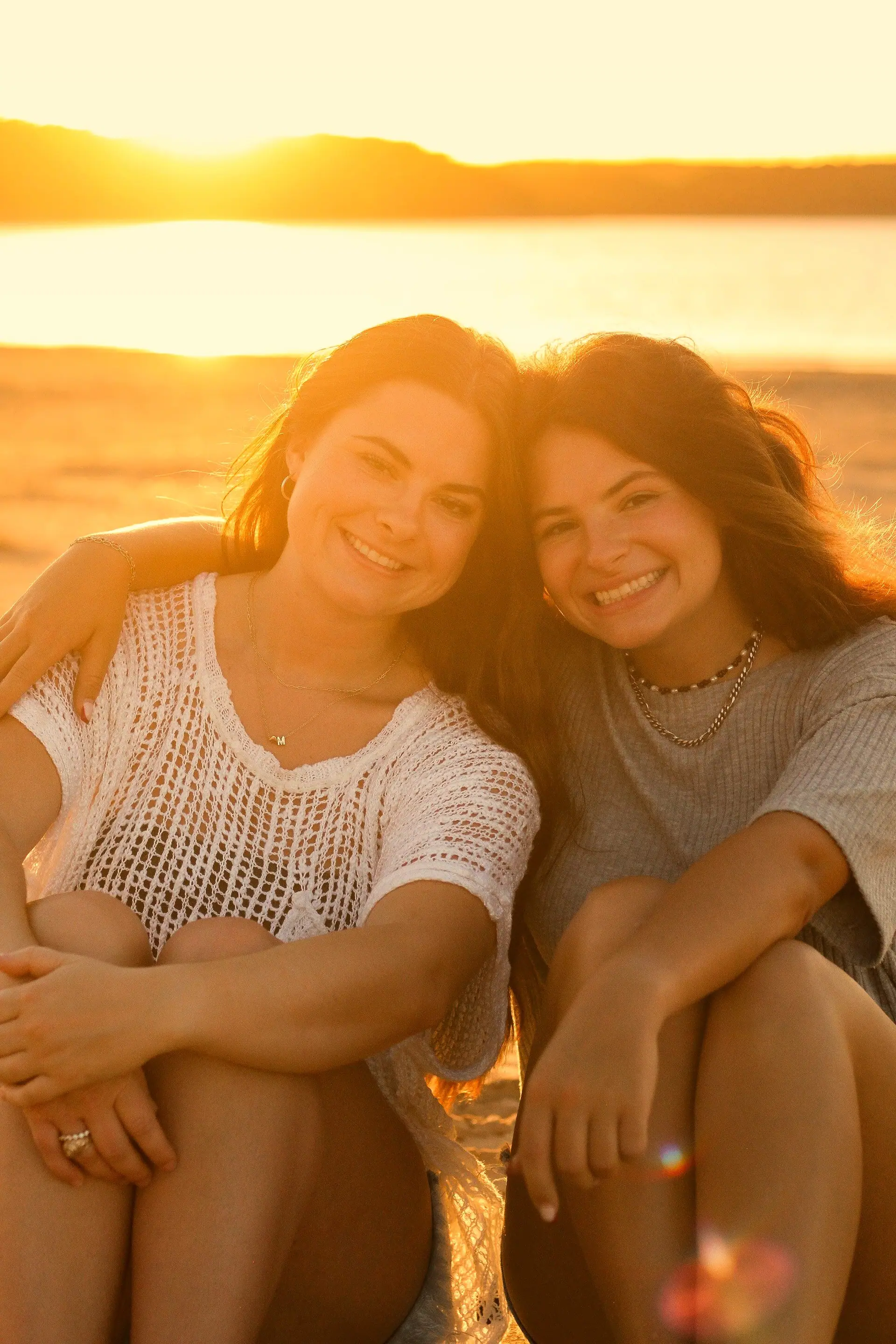 Duas amigas abraçadas sorrindo para a câmera na praia ao entardecer